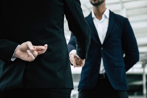 Business man crosses his fingers behind his back while shaking another business man’s hands.