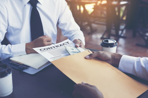 Business man hands packet containing contract and cash bribe to another man.